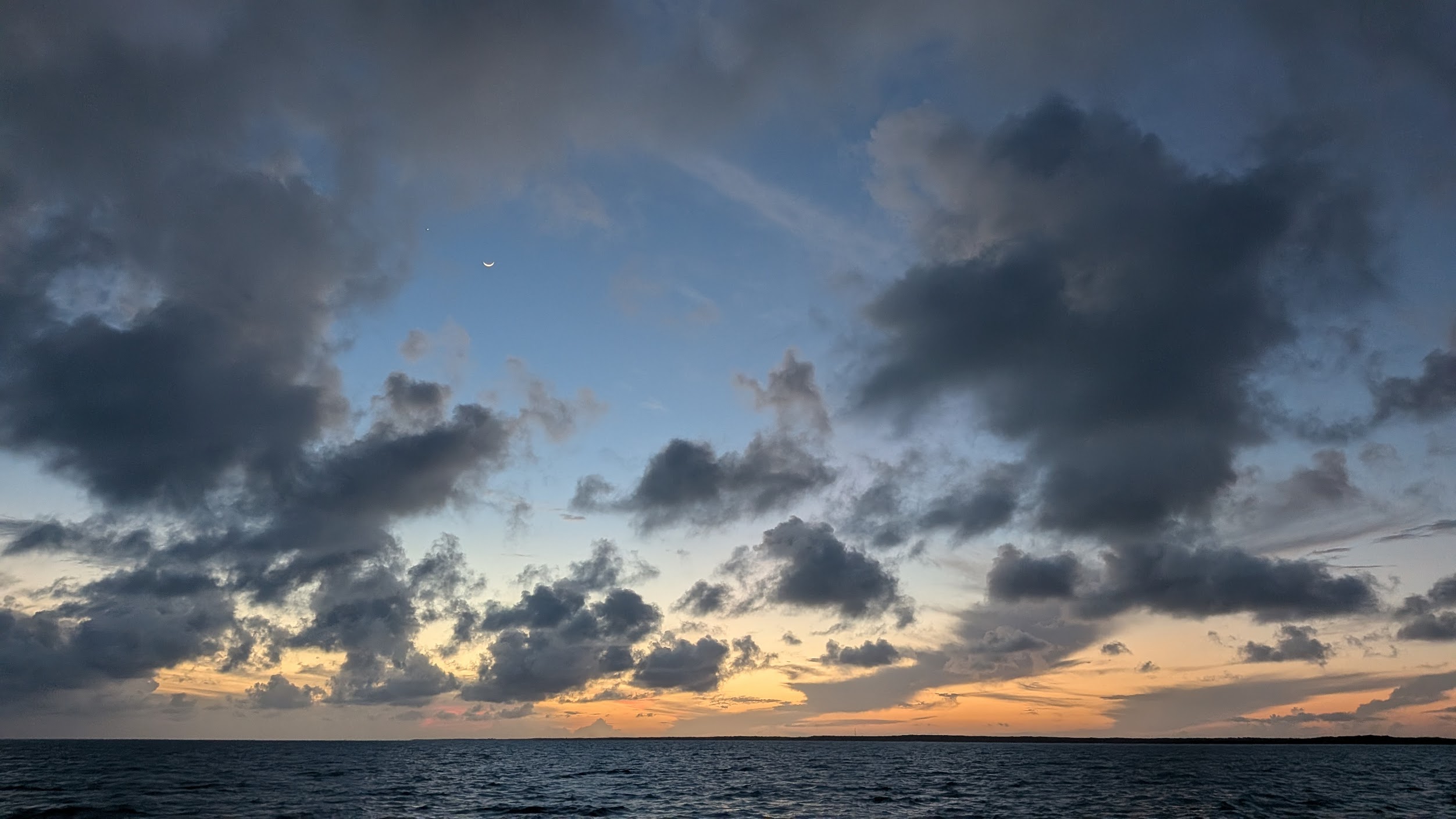 A crescent moon over scattered clouds at dusk, above open water