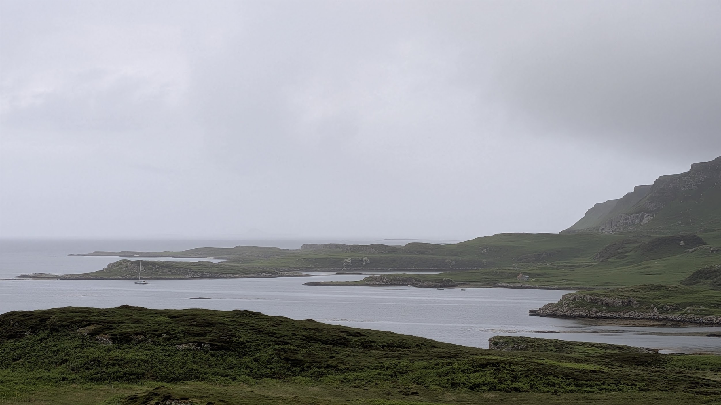 A sailboat anchored in a green-shouldered bay under low coastal cloud