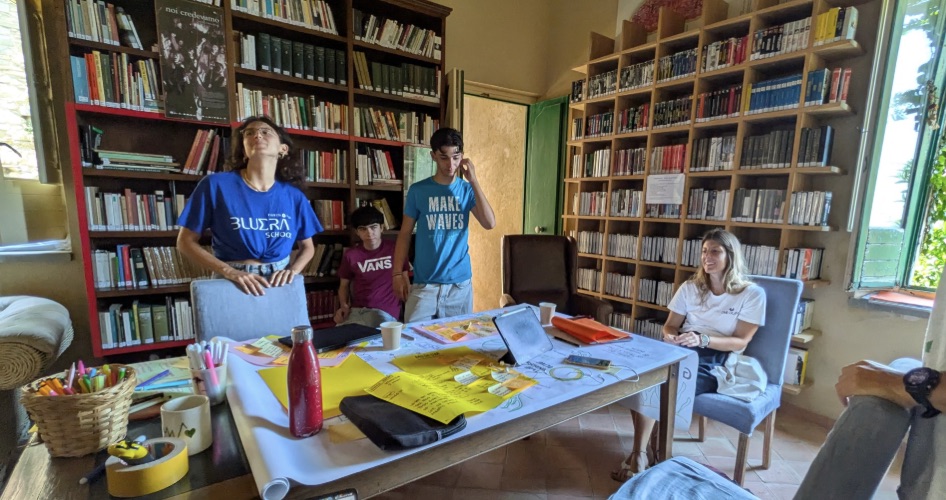 A design session around a paper-covered table in a library: markers, sticky notes, a laptop, and four people working the sheet