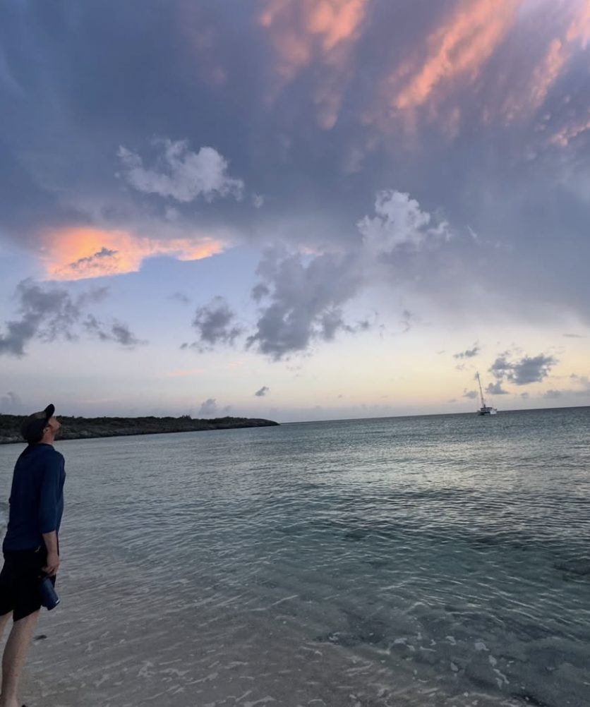 Looking up at a pink and grey dusk sky from a shallow beach, the catamaran anchored offshore