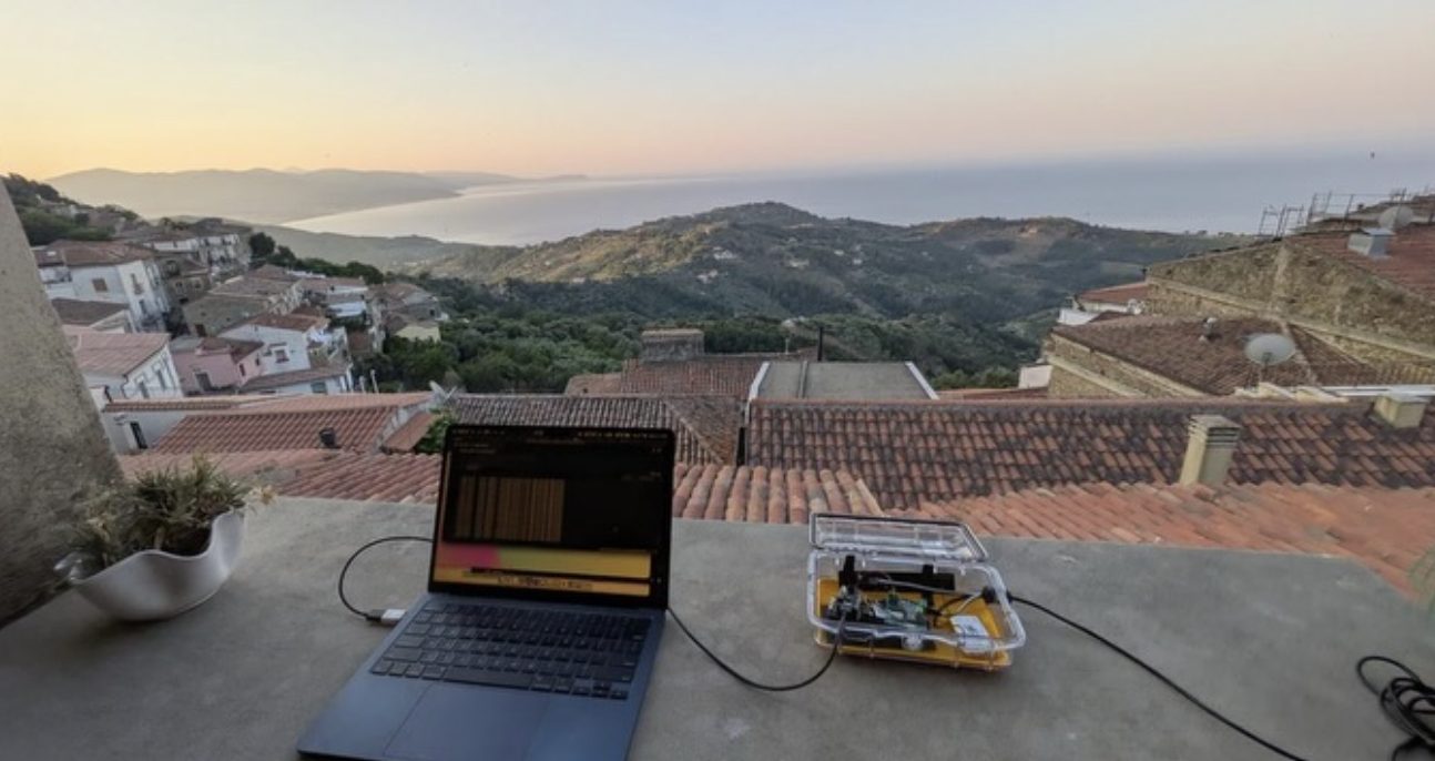 A laptop and an open sensor case on a terrace wall at dawn, tiled roofs and the bay below