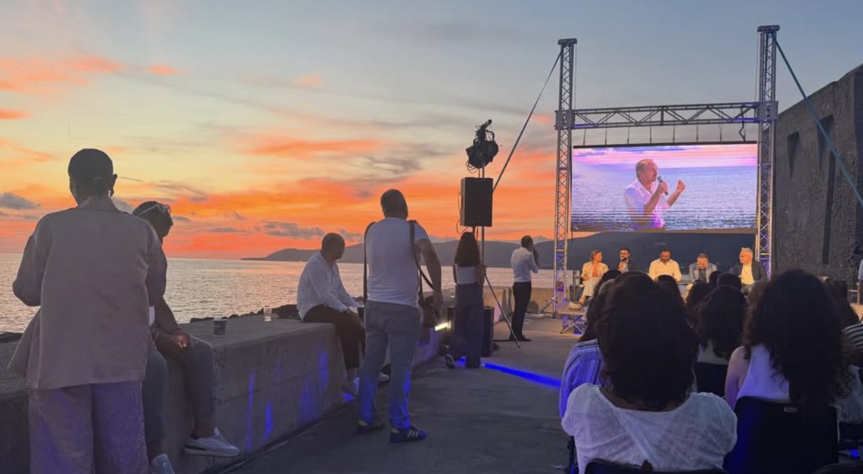 The summit stage on the harbour wall at sunset, a panel on screen, the audience facing the sea