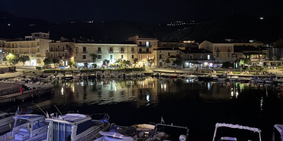 The harbour at night, boats in the foreground, lit houses on the quay reflected in still water